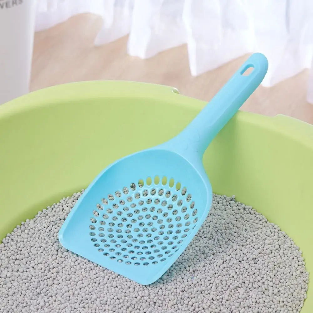 A light blue plastic cat litter scoop with sifting slots resting on a pile of gray cat litter.