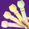A hand holds a set of pastel-colored squeeze spoons for feeding liquid cat treats against a purple background.