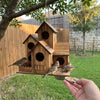 A hand holds a handcrafted wooden bird house feeder with multiple entrances, suspended from a tree branch.