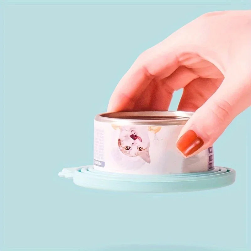 A hand is holding a light blue pet food can lid and spoon set against a pale teal background.
