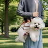 A person is holding a white fluffy dog in a breathable sling carrier while standing outdoors in a grassy park.