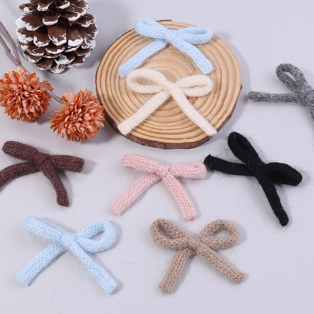 A set of six handmade knitted cotton bows in various shades of beige, pink, blue, and black arranged around a wooden slice.