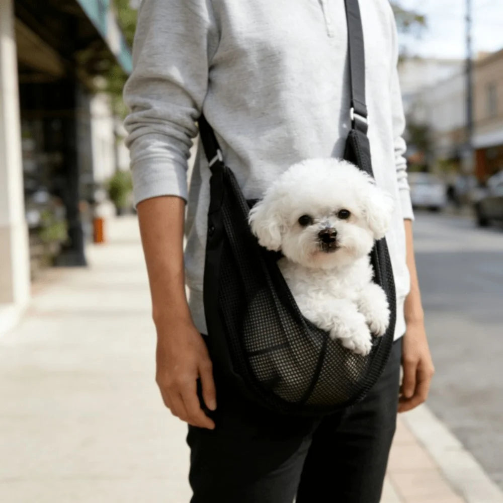 A person is carrying a black mesh pet carrier sling with a fluffy white dog in it while walking outdoors.