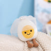 A cute white egg-shaped plush toy with a yellow yolk and small stitched smile resting on a light fabric surface.