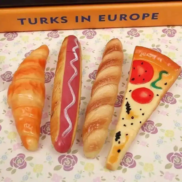 A collection of novelty food-shaped ballpoint pens, including a croissant and a hot dog, displayed on a patterned background.