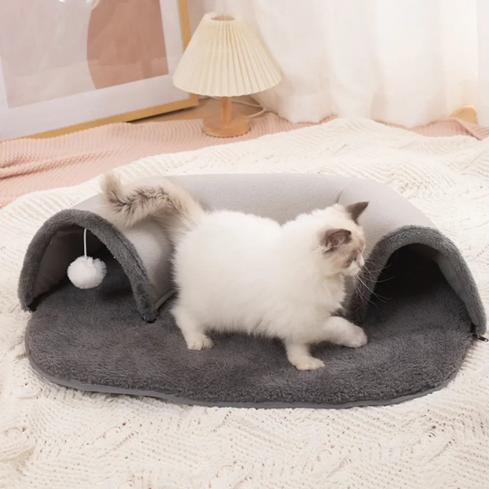 A fluffy white kitten is relaxing inside a gray oval cat tunnel and cozy bed nest on a white textured blanket.