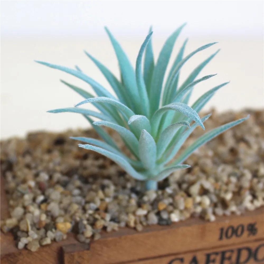 A small, blue artificial succulent plant growing in coarse gravel on a wooden base.