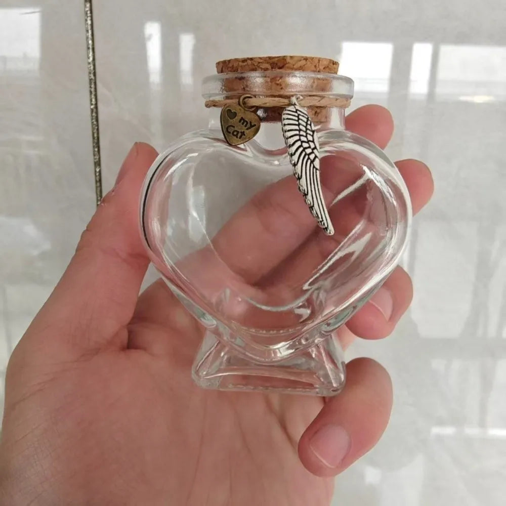 A clear glass heart-shaped jar with a cork stopper and a feather decoration is held in a hand.