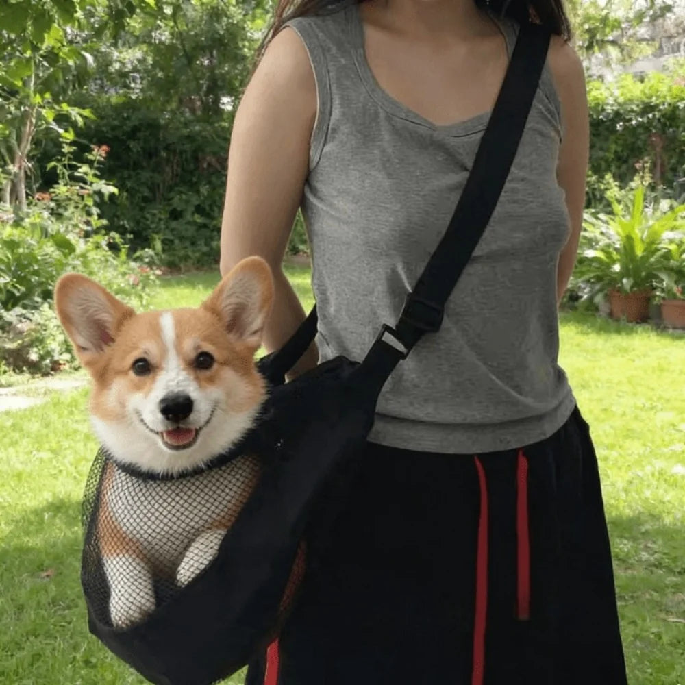 A person is wearing a black pet carrier sling with a small dog in the carrier outdoors.