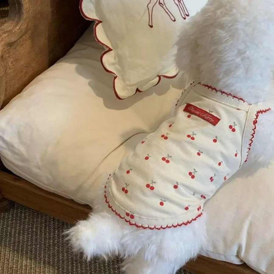 A white pet tank top with a cherry print design is shown resting on a white fabric surface.