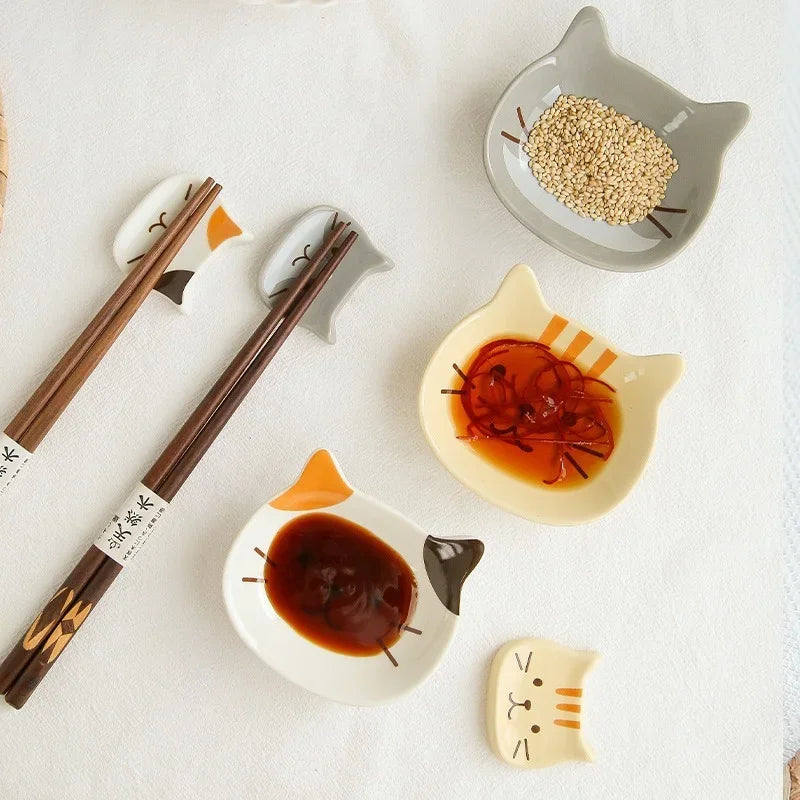 A collection of cat-shaped ceramic sauce dishes and chopsticks on a white cloth, including a dish with dark sauce and another
