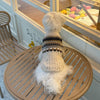 A white dog wearing a beige and black plaid knit sweater sitting on a wooden table.
