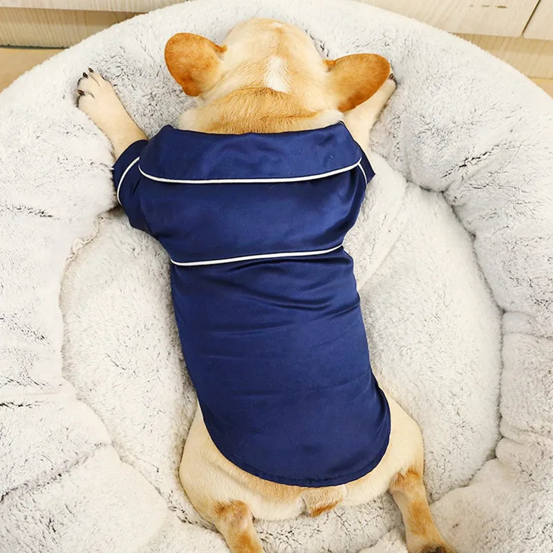 A light brown dog is lying down inside a white fluffy bed wearing a navy blue silk pet pajama top.