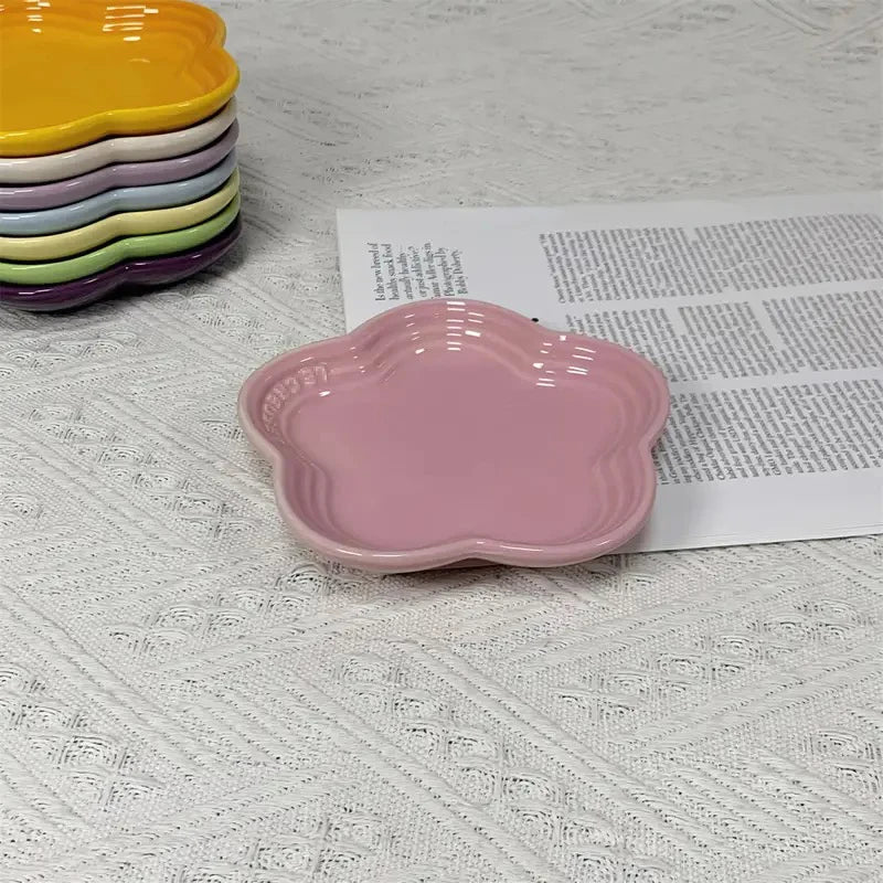 A pink flower-shaped ceramic pet bowl and dipping dish is shown next to a stack of colorful bowls on a white textured surface