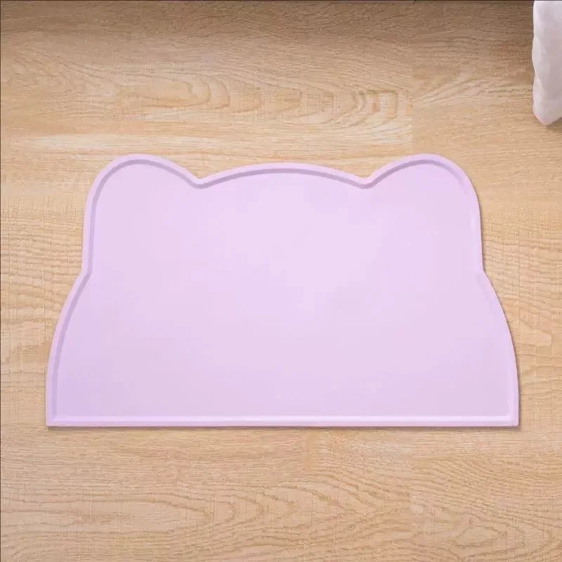 A pink silicone cat face pet feeding mat for food and water bowls placed on a wooden surface.