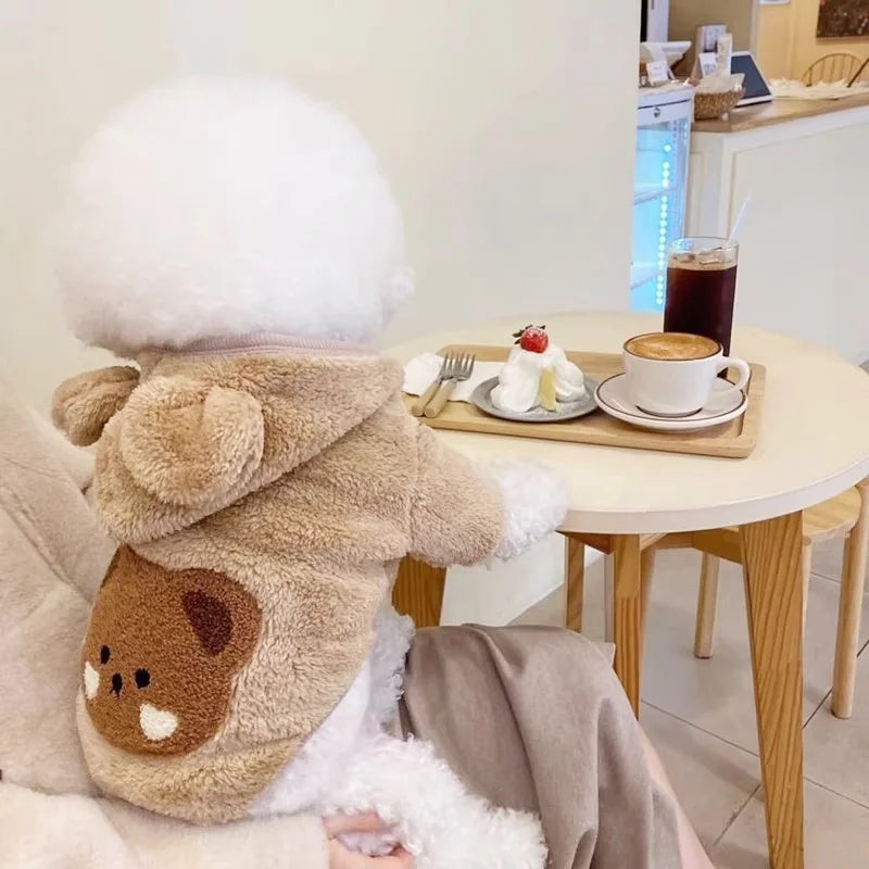 Un ours en peluche blanc et duveteux portant un sweat à capuche de chien en polaire marron, assis à côté d'une table.