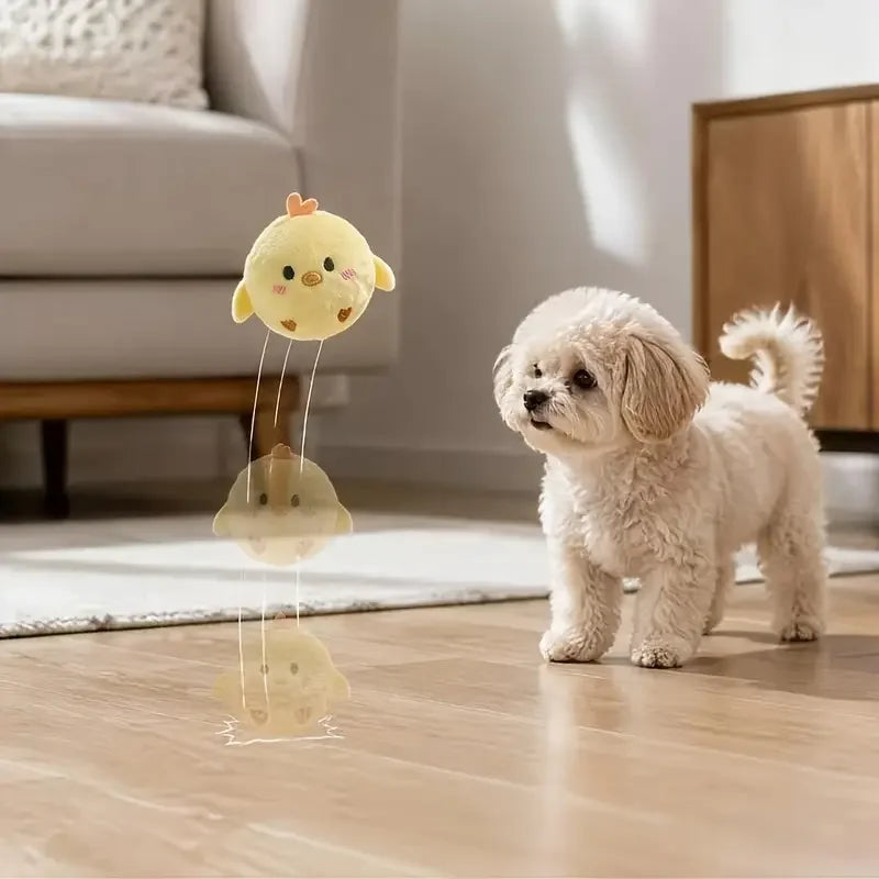 A yellow squeaky ball dog chew toy with a spiky texture is shown hanging from a string on a wooden floor.