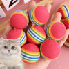 A hand holds several colorful rainbow-striped foam rattle balls, including pink and yellow, next to a small kitten.