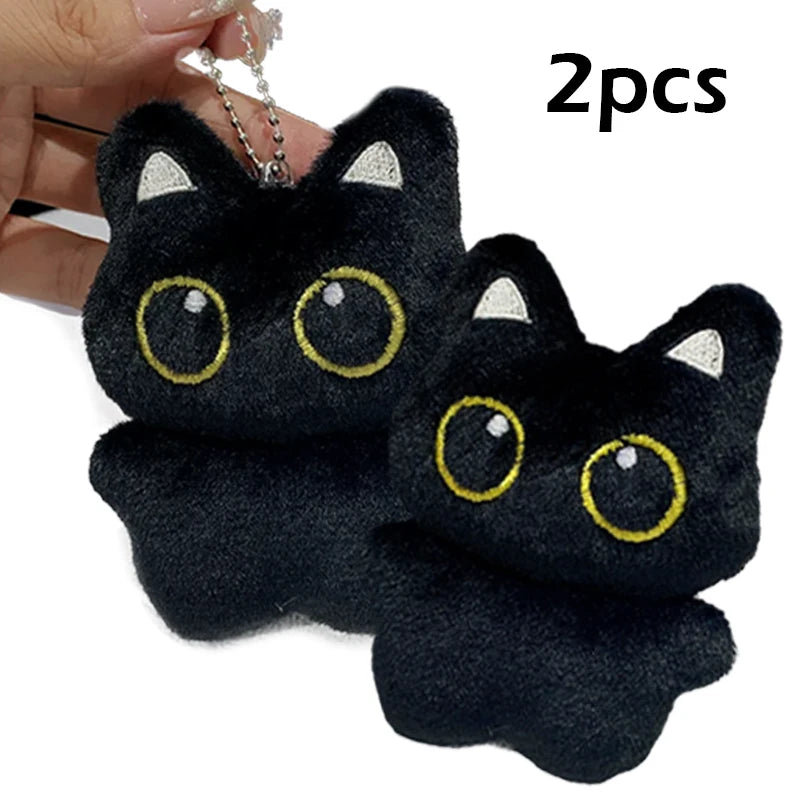 Two black cat plush pendants with gold eyes and white accents shown against a hand.