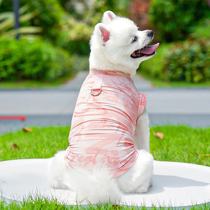 A white dog wearing a pink ice silk cooling vest sits on green grass.