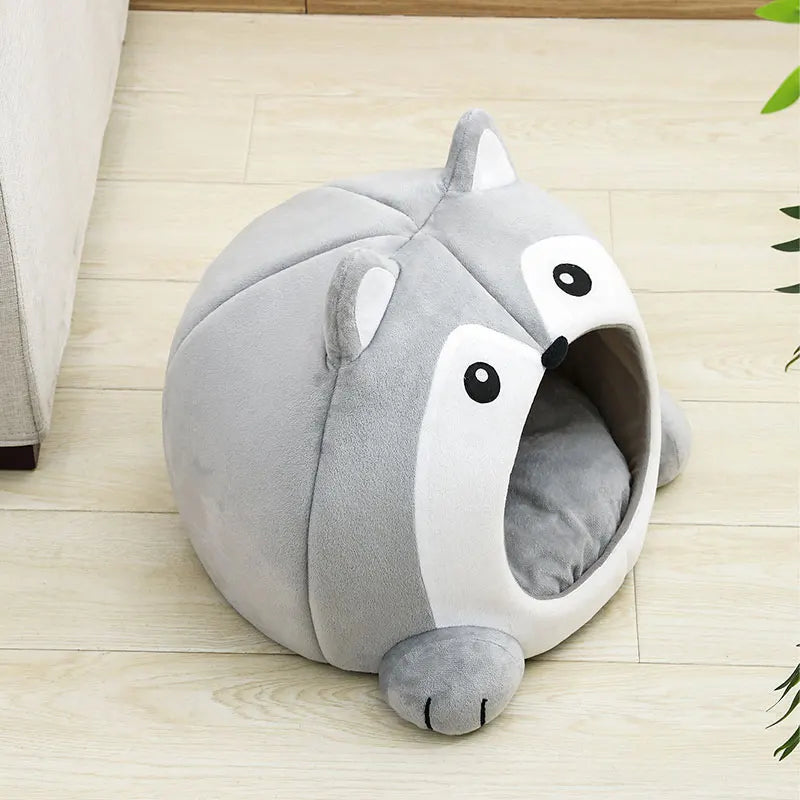Un lit pour chat en peluche gris en forme de renard avec de grands yeux et une bouche ouverte, posé sur un parquet en bois cl