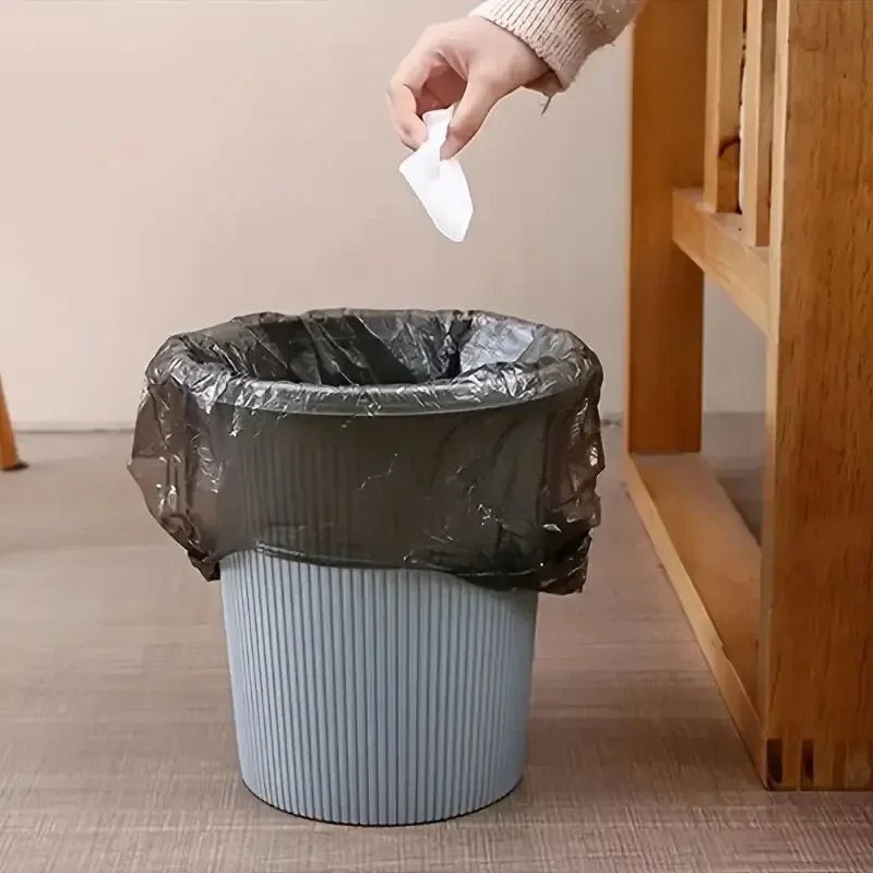 A hand is placing a white paper towel into a dark plastic garbage bag that is sitting in a light gray cylindrical container.