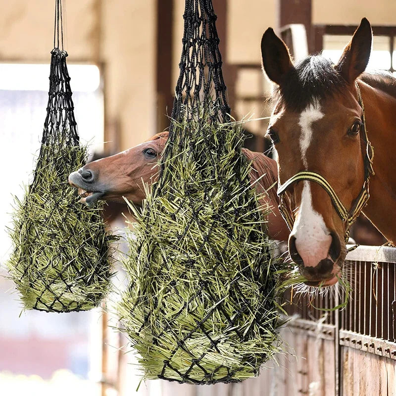 A brown horse is eating hay from a large, green, fibrous net hanging in front of a wooden fence.