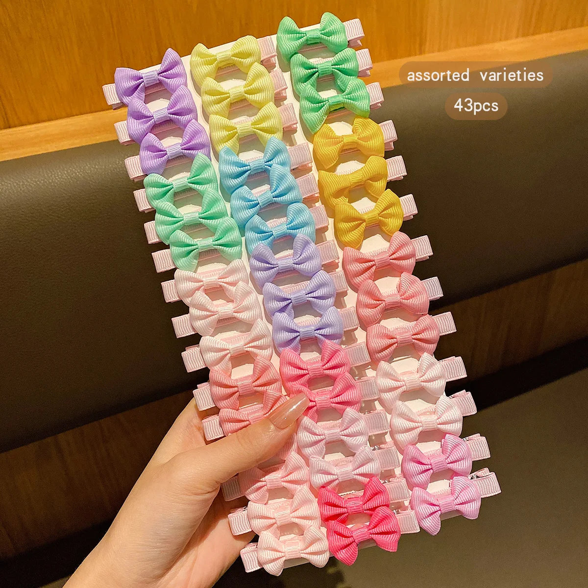 A hand holds a set of 43 assorted colorful fabric hair clips in various shades of purple, green, yellow, and pink.