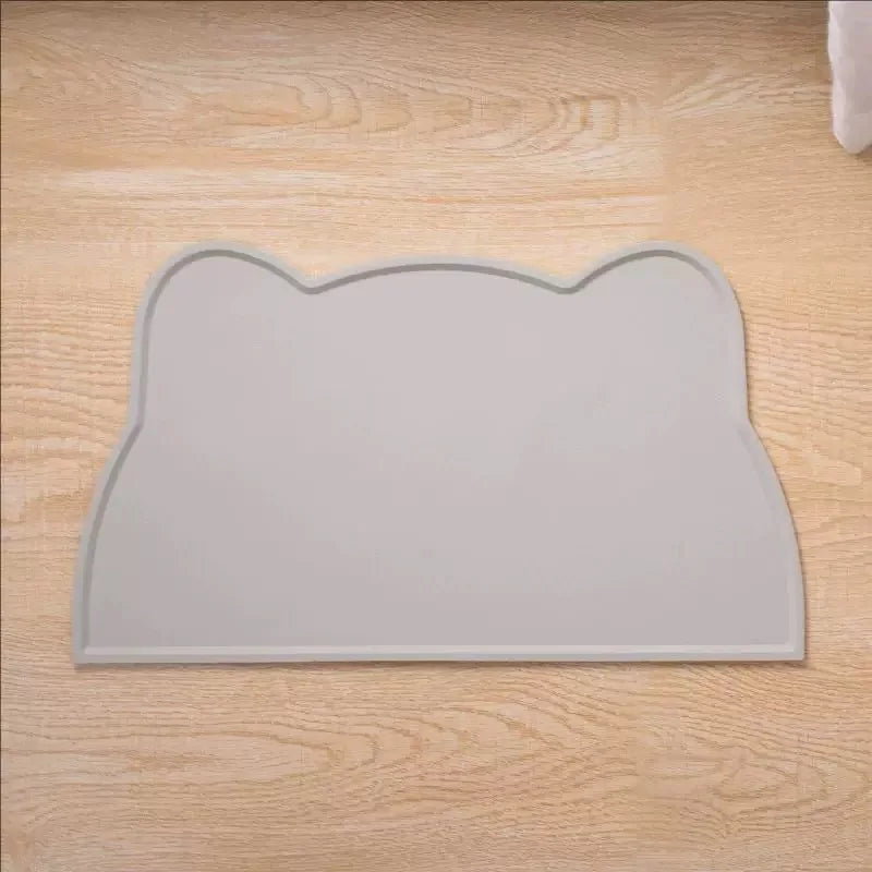 A light gray silicone cat face feeding mat is shown on a wooden surface.