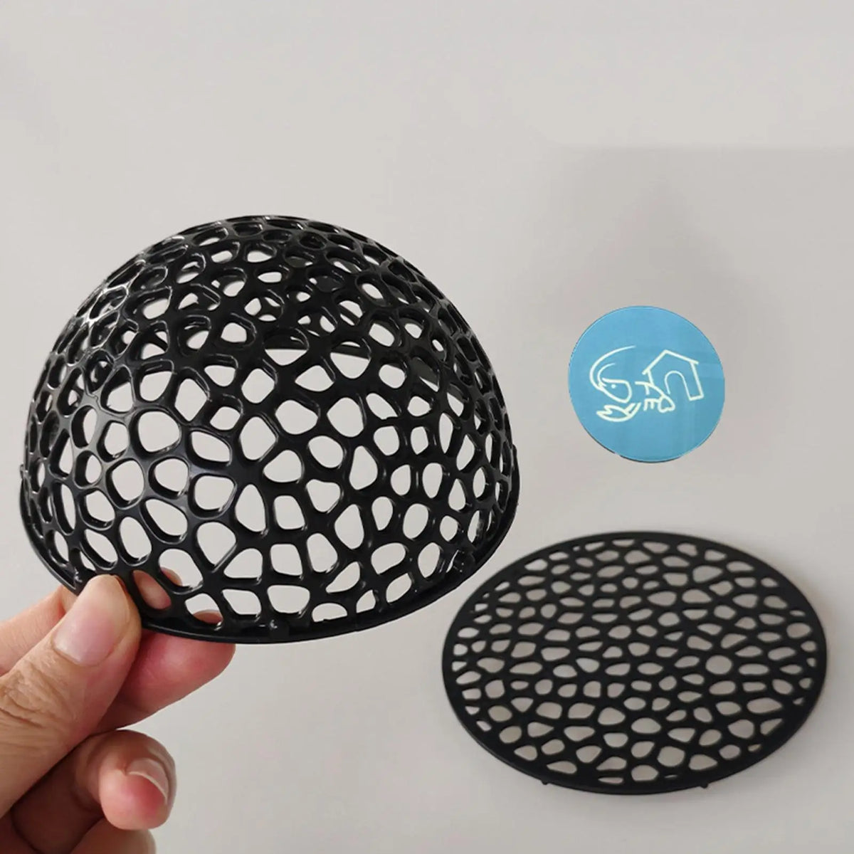 A black, perforated aquarium hideaway cave structure is shown being held by a hand next to its circular template.