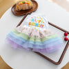 A colorful, tiered birthday outfit for a puppy featuring a white top with 'Happy Birthday' text and pastel tulle layers.