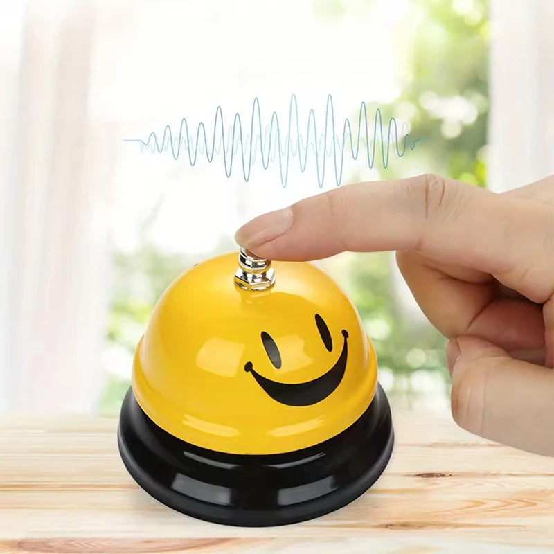 A hand is pressing a yellow classic brass call bell with a smiling face onto a black base.