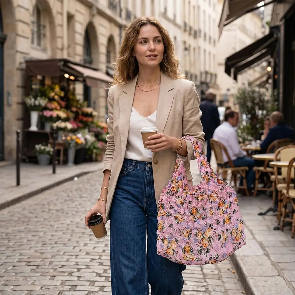 A woman wearing a beige blazer and jeans holds a floral patterned tote bag and a coffee cup on a cobblestone street.