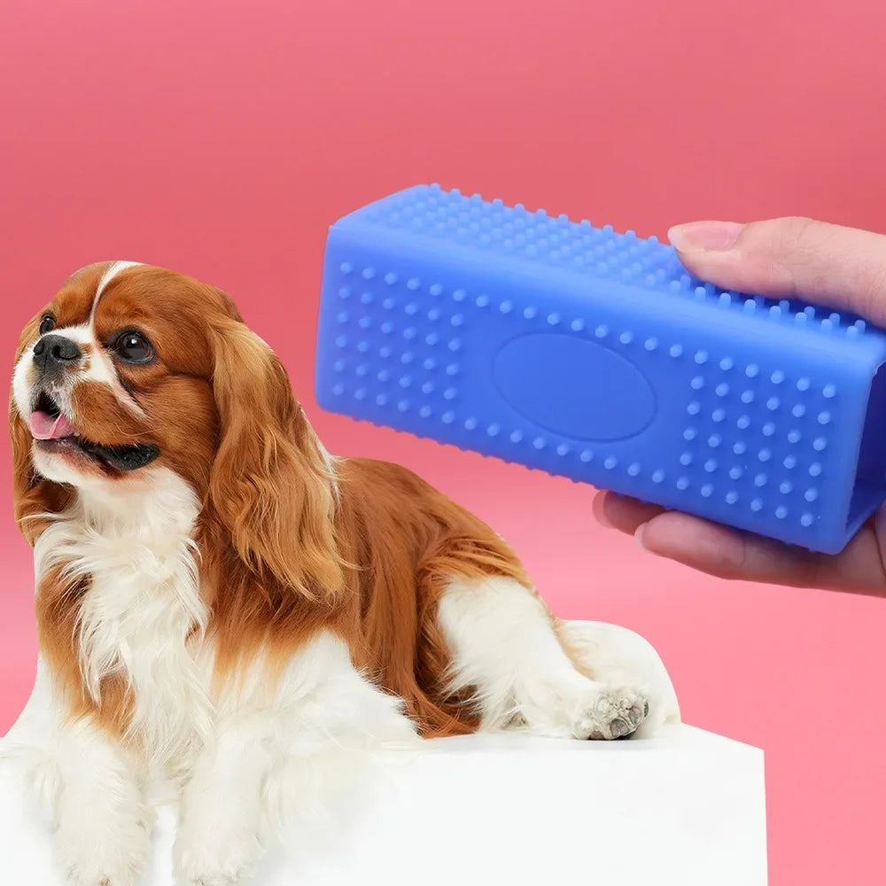 A brown and white dog is sitting next to a blue silicone pet hair remover brush being held by a hand.