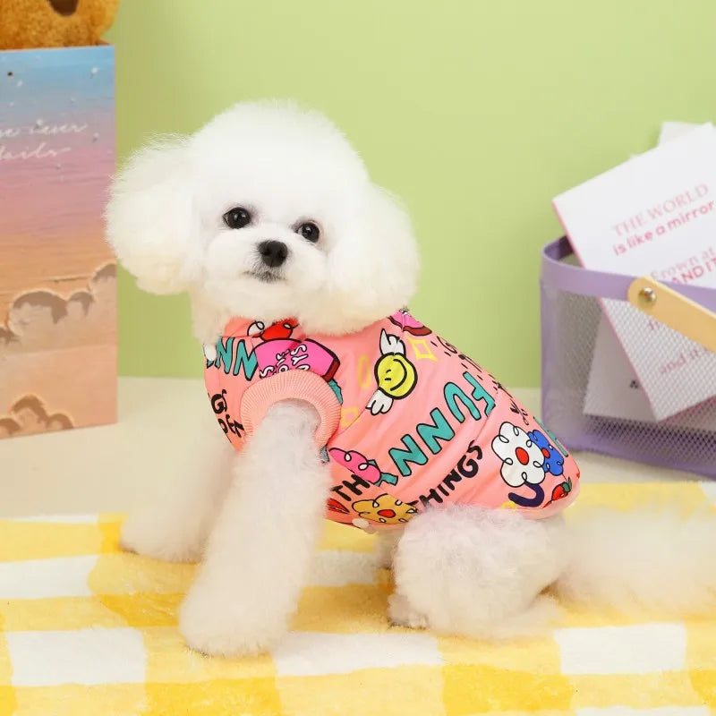 A white poodle wearing a pink and yellow patterned dog coat sitting on a yellow checkered cloth.