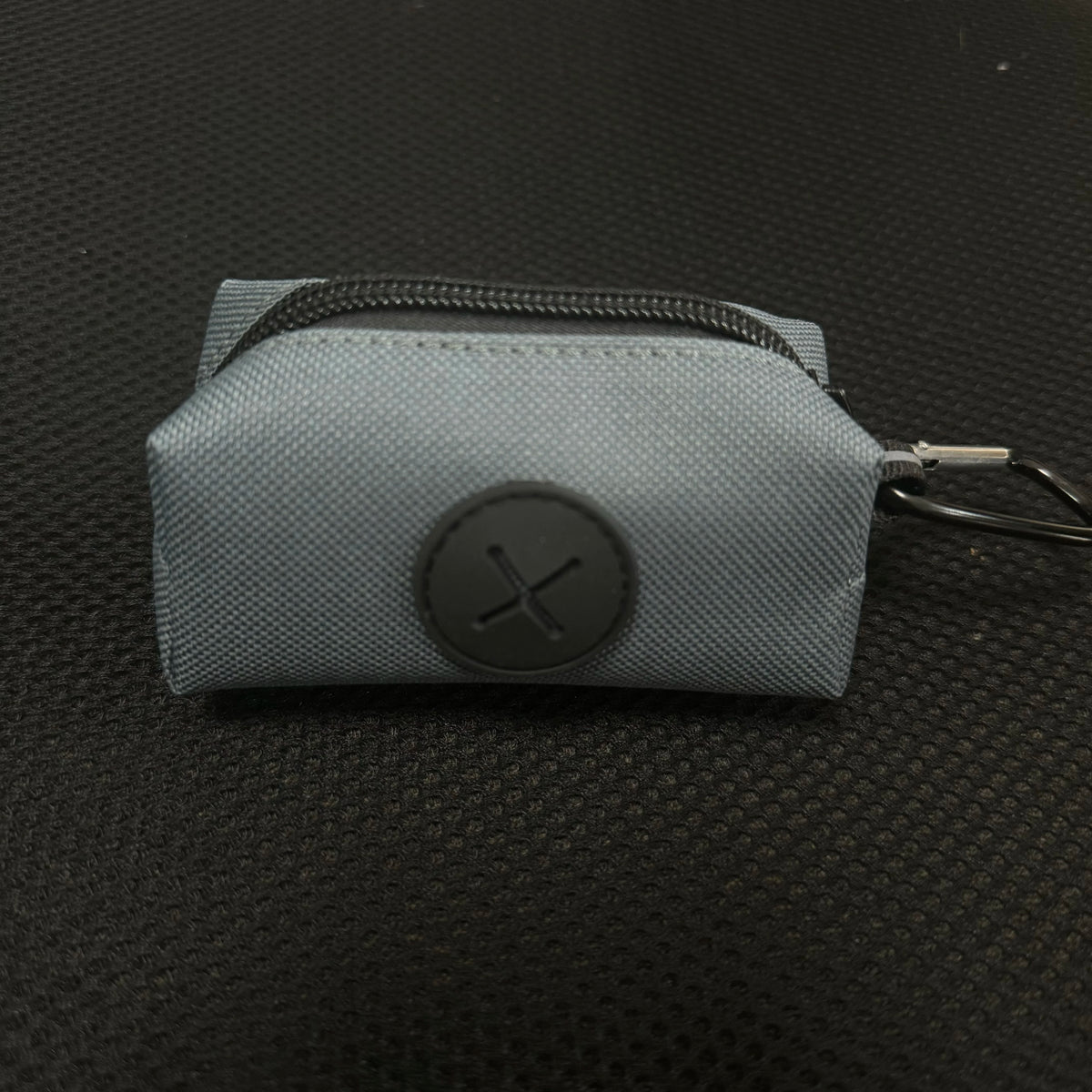 A light gray canvas pouch for a dog poop bag dispenser with a black cross emblem and a metal carabiner clip.