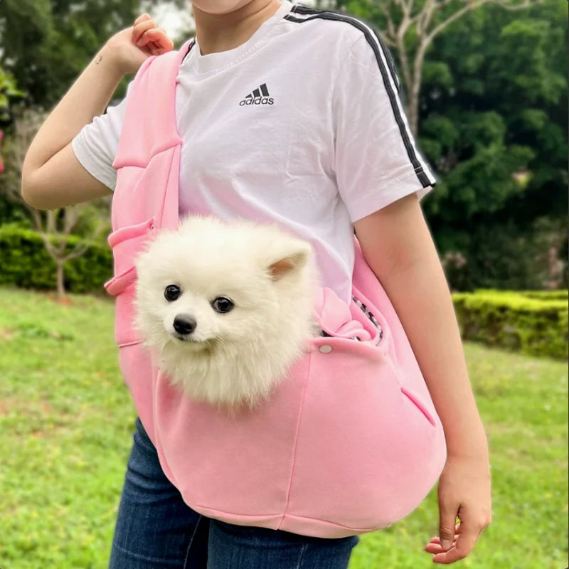 A person is holding a pink hands-free pet sling carrier with a white fluffy dog inside, outdoors.