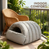 A beige semi-enclosed pet bed with a leather trim sits on a balcony railing next to a wicker chair and greenery.