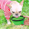 A light-colored French Bulldog is licking a green silicone collapsible pet travel bowl held by a person's hand.