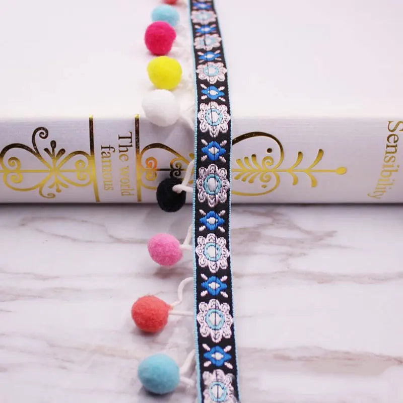 A close-up of a black lace trim featuring colorful pom poms and beads, displayed next to a rolled paper.
