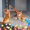 A ginger cat is playing with colorful plush balls launched by a blue toy launcher.