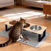 A tabby cat is sitting on a gray feeding mat next to an elevated wooden bowl stand with stainless steel bowls.
