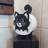 A black cat figurine with a tail sitting on a white bathroom roll holder.