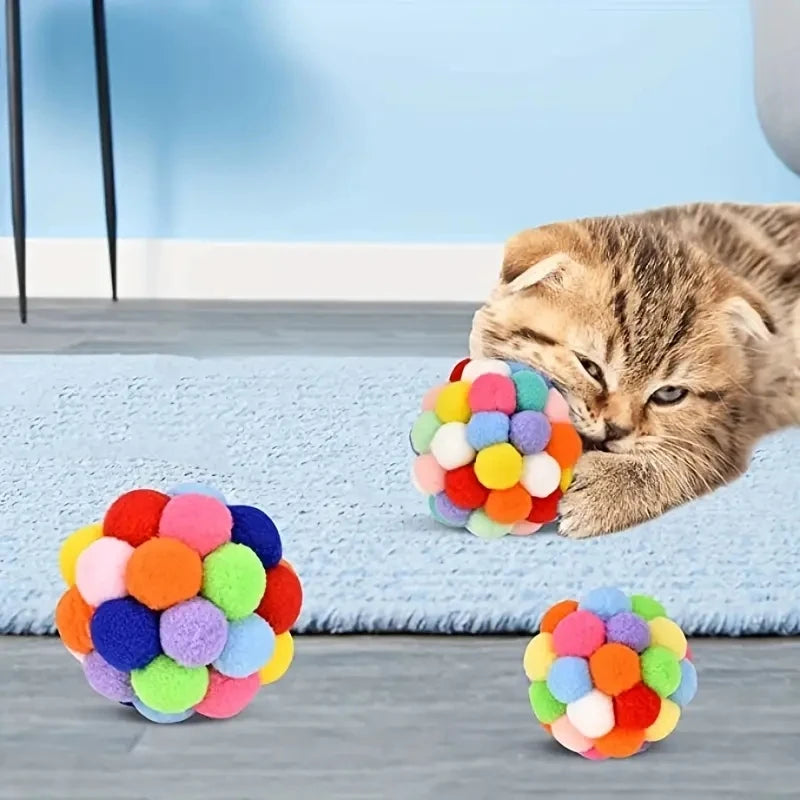 Eine tabbyartige Katze interagiert mit einem farbenfrohen, mehrtexturierten Ballspielzeugset auf einer hellblauen Oberfläche.
