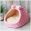 A pink, polka-dotted cozy cave pet bed with a fluffy interior is shown on a light wooden surface.