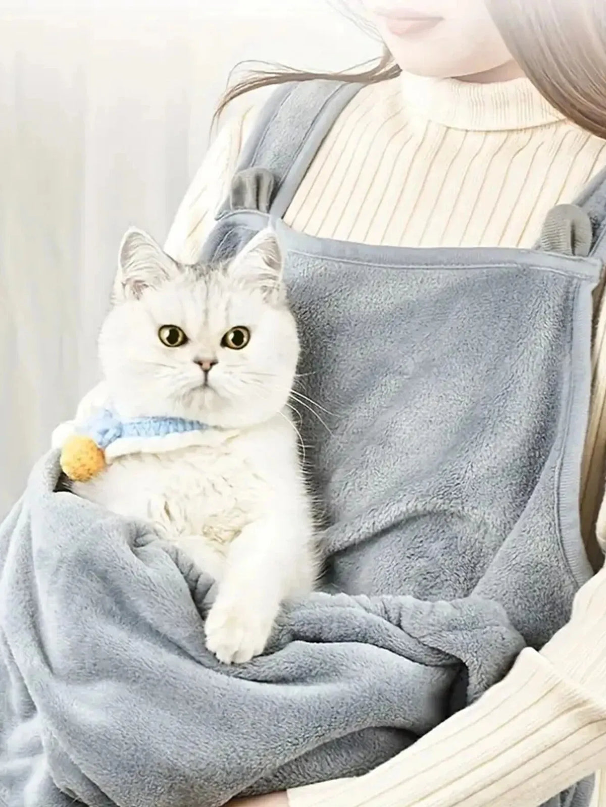Un chat blanc et duveteux se repose dans un tablier de transport pour animaux gris et confortable porté par une personne.