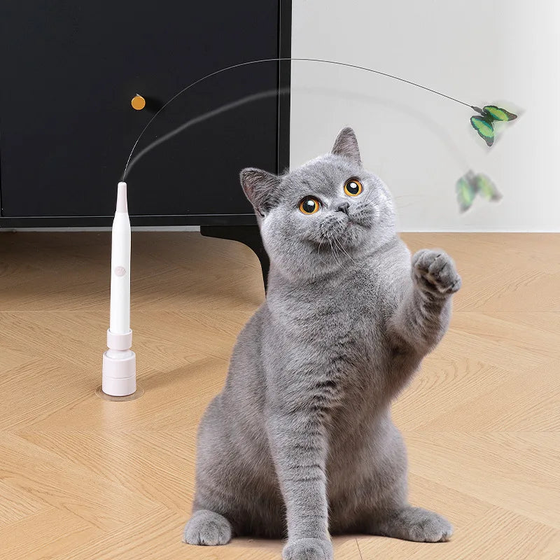 A gray cat is sitting while an electric rotating butterfly toy is aimed at it with a thin wire.