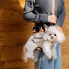 A person is holding a white fluffy dog in a beige breathable sling carrier while standing indoors.