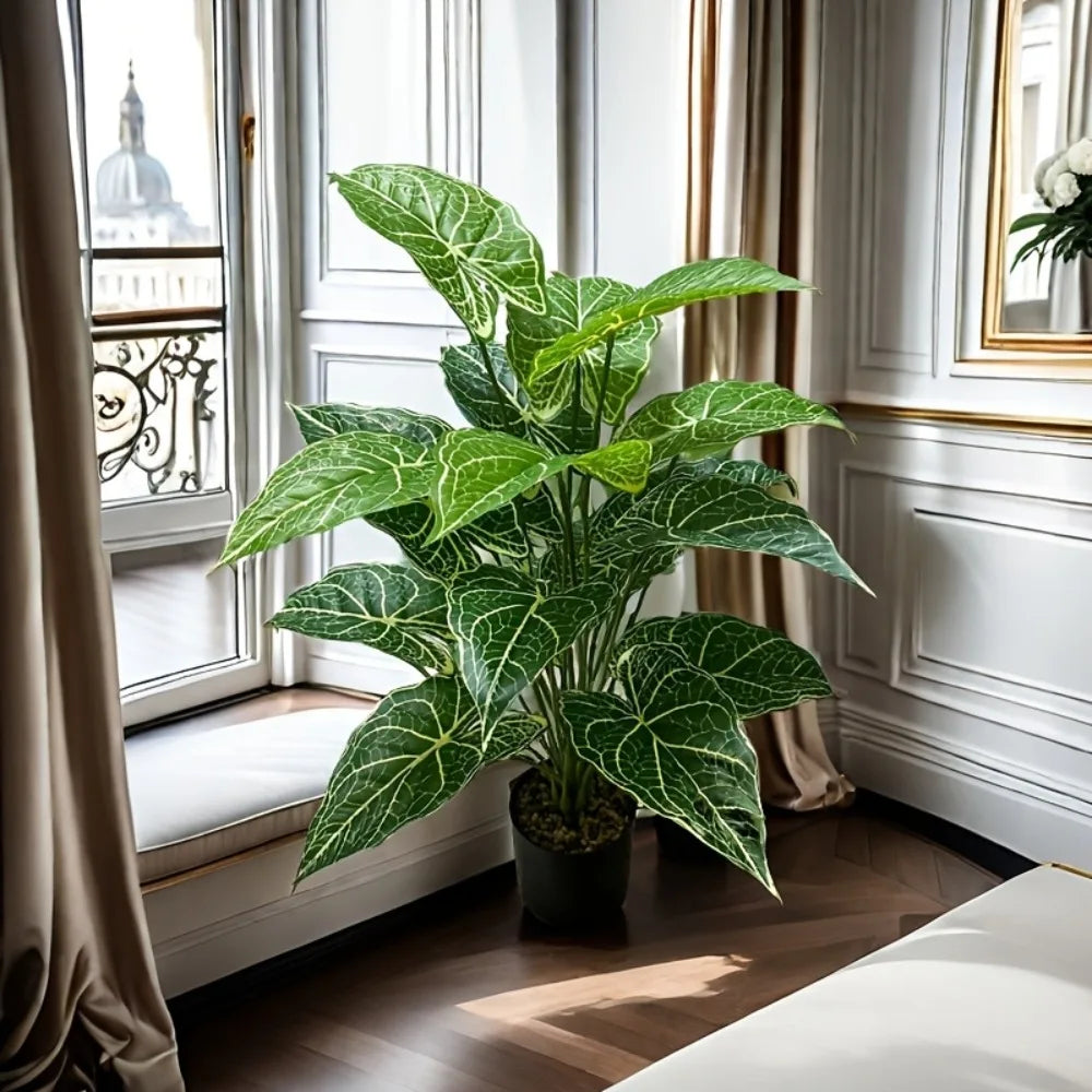 A large artificial monstera plant with green patterned leaves sits in a pot on a wooden floor next to a window.