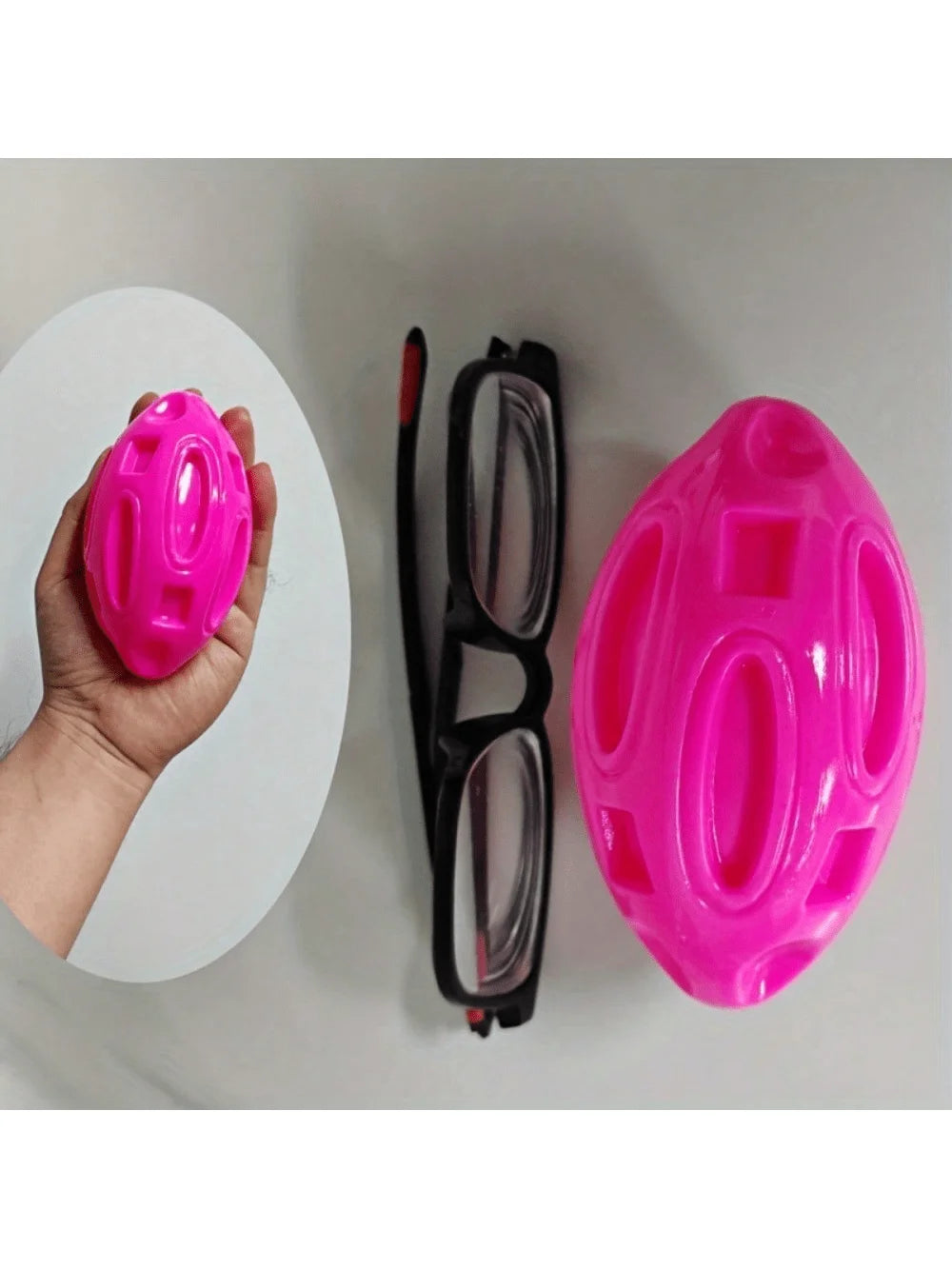 A hand holds a bright pink indestructible squeaky rugby ball dog chew toy next to a pair of black glasses.